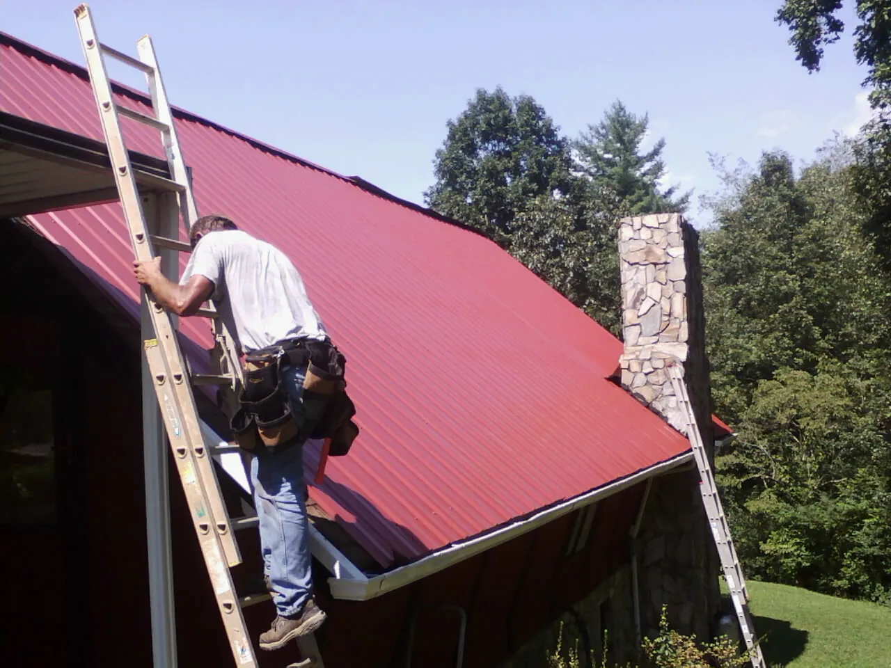 Roofer on a ladder beside a red metal roof and stone chimney — Soffit & Fascia Repair by Roofing Brothers in Reevesville